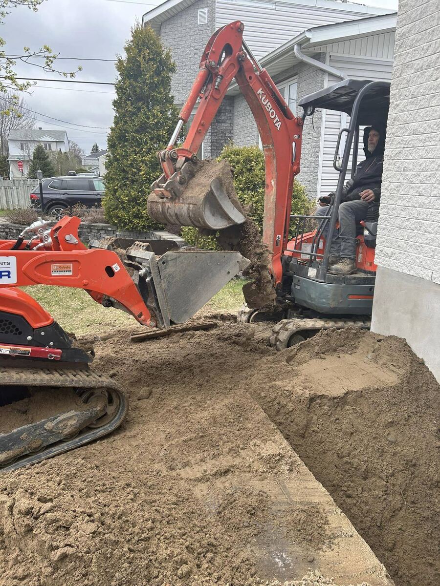 Mini-pelle en action entre une maison et une haie